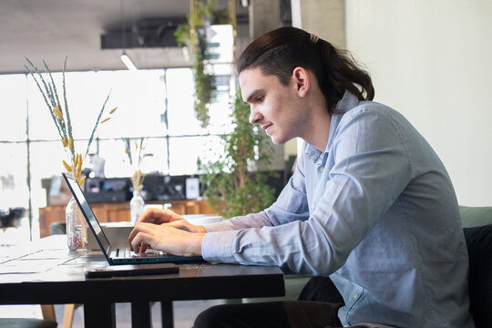 A young man sits at a cafe table focused on his laptop while sipping coffee.