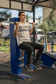 A young man works out in an outdoor gym located in a city park. He uses a pull-up bar to build strength.