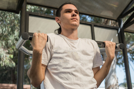 A young man works out in an outdoor gym located in a city park. He uses a pull-up bar to build strength.