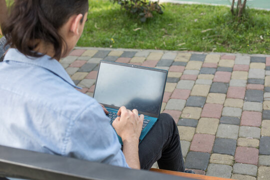 A man sits on a bench in a city park using his laptop. He has a bicycle leaning against the bench and is focused on his work