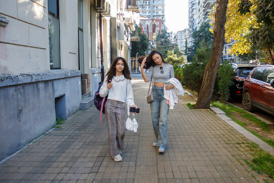 Two people walk on a city sidewalk in the afternoon with buildings and trees in the background