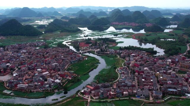 An aerial view of a rural settlement with dense buildings, winding rivers, and karst peaks in the distance.