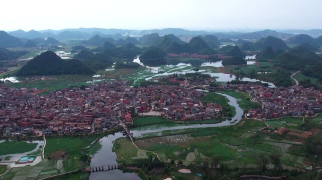 Aerial panorama of a riverside settlement nestled among karst mountains and wetlands, with dense buildings and winding waterways.