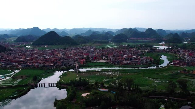 Aerial view of a riverside village with red-roofed houses, karst peaks and green farmlands.