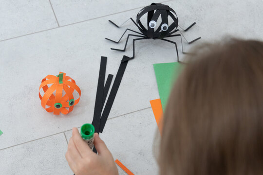 Child Making Halloween Paper Spider And Pumpkin Craft On Light Tile Floor With Marker, Using Colorful Strips, Googly Eyes, And Handmade Shapes For Festive Seasonal Art, Classroom Activity