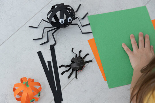 Halloween Craft Workspace With Paper Spiders, Black Cut Shapes, Orange Decorative Accent And Hand Arranging Bright Green And Orange Sheets On Gray Surface, Showing Seasonal Diy Decoration