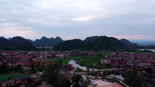 A rural settlement sits along a winding river, backed by karst mountains under an overcast sky, with lush green fields surrounding it.