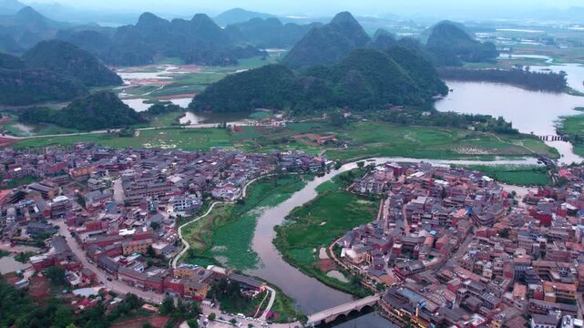 Aerial view of a riverside settlement nestled among karst mountains and green wetlands, with dense buildings and natural landscapes.