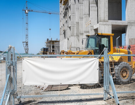 An empty white advertising banner hangs on a metal gate at a building site. A large yellow heavy machinery loader stands near a high concrete structure, while the construction site also features an in
