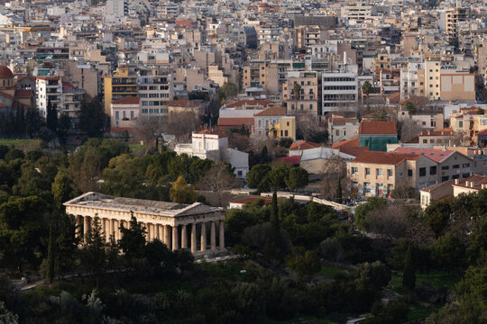 Aerial view of the well-preserved ancient Temple of Hephaestus nestled in the lush Ancient Agora park, contrasting with the dense, modern residential rooftops of Athens, Greece.