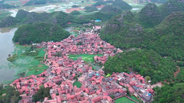 An aerial shot shows a traditional village with red rooftops nestled among lush green mountains and water bodies.