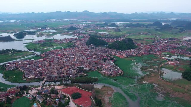 High-angle aerial panorama shows a dense village cluster surrounded by wetland, karst hills and water networks, with distant mountains.