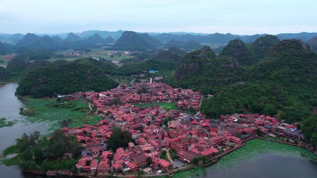A traditional village with red roof buildings nestled among green karst mountains and water bodies, forming a serene rural landscape.