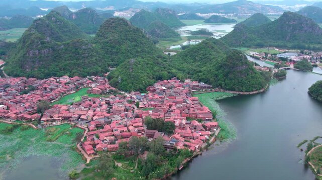 An aerial view of a traditional village with red-roofed houses, surrounded by green mountains and calm water bodies.