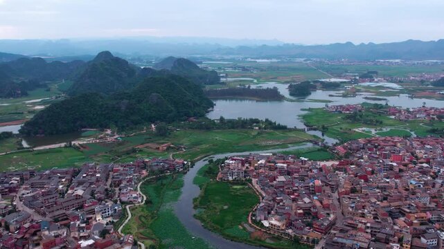 An aerial panorama shows a waterside town nestled among forested mountains and winding lakes in a rural landscape.