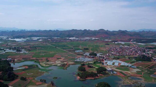 An aerial shot shows a rural landscape with farmland, water bodies, villages, and distant mountain ranges under a clear sky.