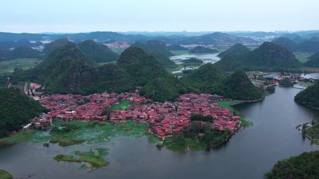 Aerial view of a red-roofed village nestled among karst mountains and calm water bodies, with lush green surroundings.