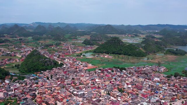 Elevated daytime view of a dense rural village nestled among karst hills and green farmlands under a clear sky.