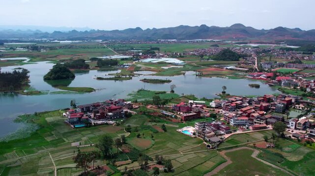 A high-angle landscape shows a lakeside village, farmlands, and distant mountains under a clear sky.
