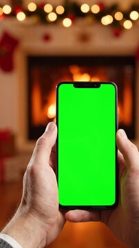 Hands Holding Smartphone With Chroma Key Green Screen Display In Front Of A Cozy Christmas Fireplace Background