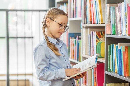 Schoolgirl reading book while browsing bookshelves in library
