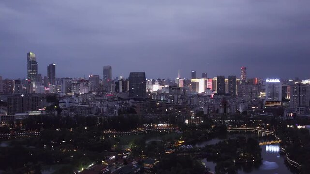 A wide aerial view of a city at dusk, featuring illuminated buildings and a park with water in the foreground.