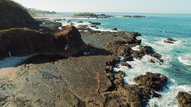 Person standing on rocky cliff by the ocean in Costa Rica. Turquoise waves crashing against dark shore of San Juanillo beach.