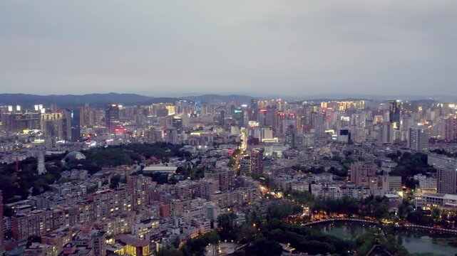 Aerial panorama of a sprawling urban city at dusk, featuring mixed buildings, water bodies, and green areas under an overcast sky.