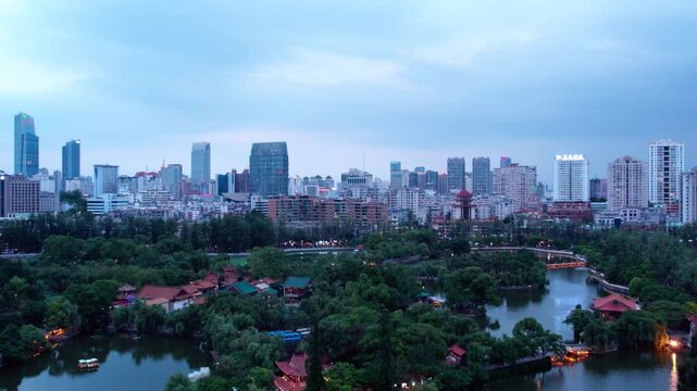 A serene city park with a lake, blending natural greenery and traditional structures, set against an urban skyline at dusk.