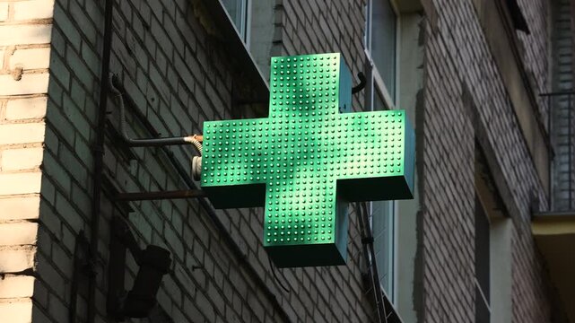 Illuminated green pharmacy cross glows on brick building exterior wall