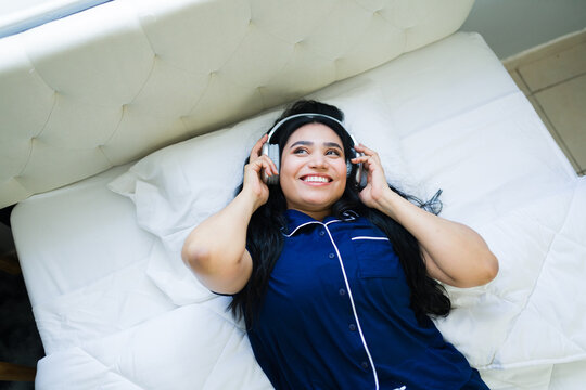 Happy young woman in pajamas lying on bed wearing headphones, enjoying music and relaxing at home