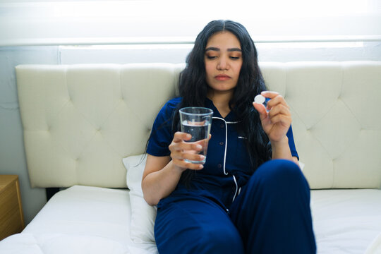 Sick woman taking medication in bed feeling unwell, holding a pill and glass of water for her health treatment