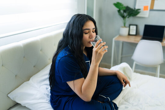 Woman in pajamas drinking glass of water in bed, practicing a healthy morning routine for hydration and wellness