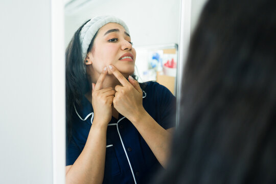 Young woman checking her skin in the mirror, looking at a pimple on her cheek and expressing frustration