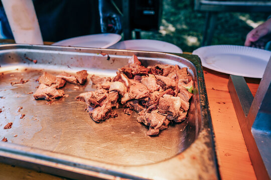 Cooked beef chunks served on a stainless steel tray at an outdoor food event with disposable plates visible in the background