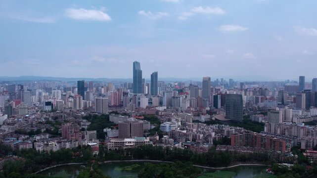A wide aerial panorama shows a sprawling urban city with dense buildings, skyscrapers, and patches of greenery under a clear sky.