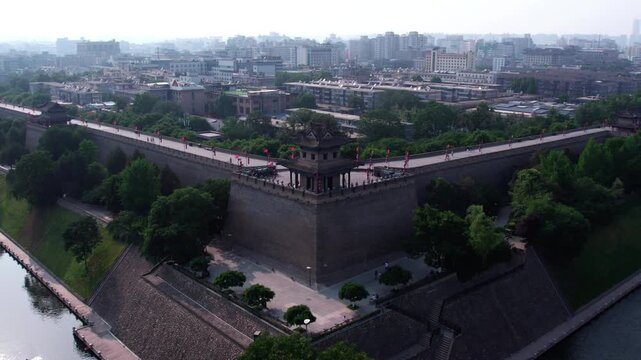 A historic ancient city wall with a corner tower, surrounded by a moat and green trees, with modern urban buildings in the distance.