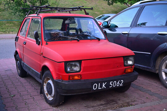 A red compact mini vintage car, a Polski Fiat 126p, parked on the street with a roof rack and worn license plate.