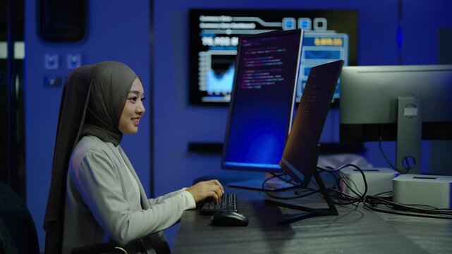 Smiling Muslim female software developer wearing a hijab typing on her computer keyboard, coding on multiple monitors in a modern dark high tech office. IT security programmer analyzing digital data.