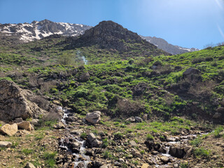 Sun-drenched green hillside rises toward snow-capped peaks, thin smoke drifting skyward beside a flowing stream under a brilliant blue spring sky. 📍GHallakhane, Daraki, Kurdistan  © Hawraman83