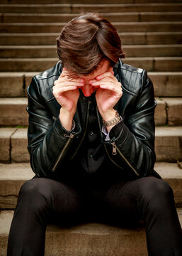A young man in a leather jacket sits alone on stone steps with his hands over his face, appearing to be in a moment of deep distress, exhaustion, or profound sadness.