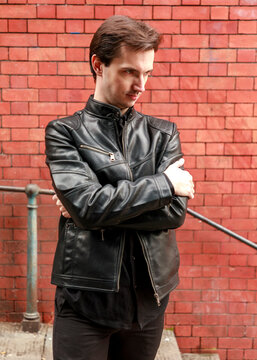 A young man dressed in a black leather jacket and matching shirt poses confidently with his arms crossed in front of a textured red brick wall during a professional fashion portrait session.