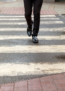 A person in formal black trousers and polished dress shoes walks across a pedestrian crosswalk in the city, suggesting a professional commute or business-related activity.