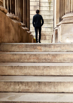 A lone man in dark clothing walks up a set of stone steps framed by massive architectural columns, moving toward a grand building entrance.