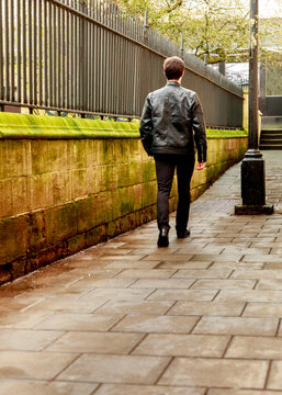 A man dressed in a black leather jacket walks away from the camera along a historic stone path beside a tall metal fence on a quiet day.