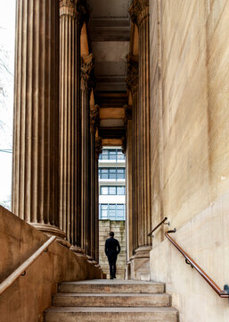 A man in a black suit walks up stairs between grand neoclassical columns, moving from traditional architecture toward a modern building, highlighting between the historic and the contemporary
