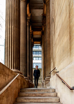 A man in black walks down stone steps between massive classical columns, creating a striking contrast between historical architecture and a modern urban environment.
