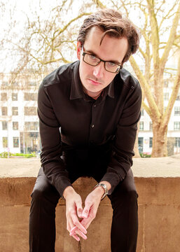 A man in a black button-down shirt and glasses sits on a stone wall, looking directly at the camera with a focused expression for a professional outdoor portrait.