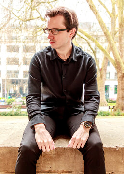 A young man wearing a black shirt and glasses poses thoughtfully while sitting on a stone ledge in a park, capturing a contemplative moment during an outdoor portrait session.