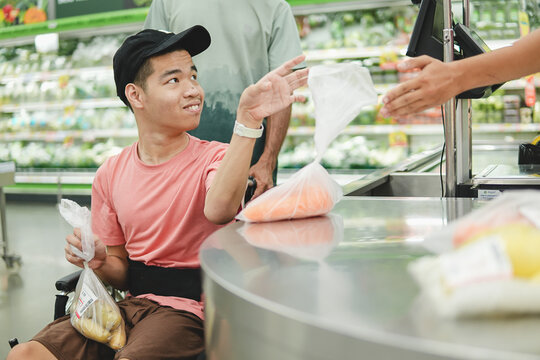 Happy young Asian man in a wheelchair handing a bag of fresh vegetables to a supermarket worker at the counter, concept of inclusive lifestyle and accessibility.